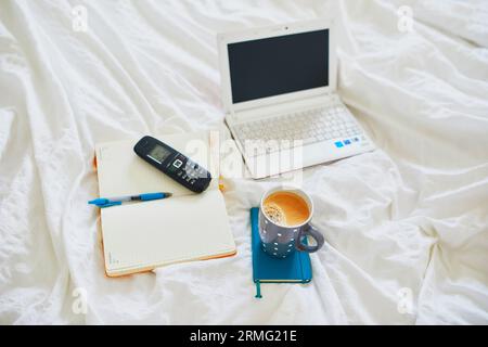 Laptop, cup of coffee, notebook and phone in bed on clean white linens. Freelance, distance learning or work from home concept Stock Photo