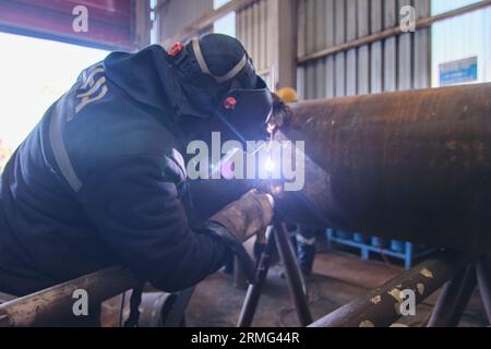 a man in full safety gear like a helmet, visor, and vest. doing welding on a rusted metal pipe. a man using a handheld electrical saw on a metal pipe. Stock Photo