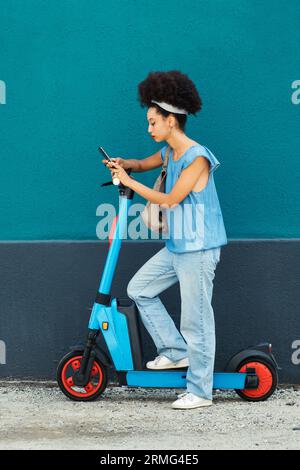 Side view of young African American female with headband on Afro hair looking at screen of mobile phone while standing with leg on electric scooter an Stock Photo