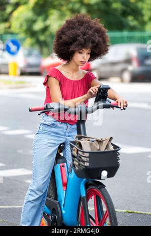 Stylish young mixed race woman with dark Afro hair in casual outfit using smartphone while riding electric bicycle on asphalt road in city on sunny da Stock Photo