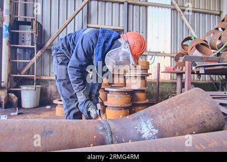 a man in full safety gear like a helmet, visor, and vest. doing welding on a rusted metal pipe. a man using a handheld electrical saw on a metal pipe. Stock Photo