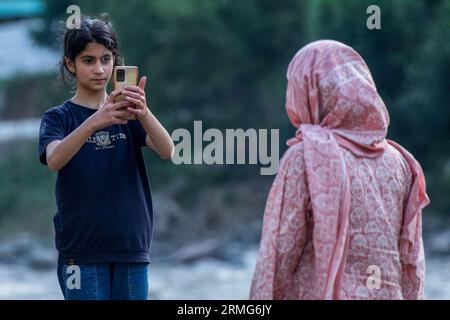 Keran Kupwara, India. 24th Aug, 2023. A girl takes a photo of her mother on the banks of Neelam river or Kishan Ganga that has divided Kashmir into two parts controlled by nuclear rivals India and Pakistan. The river acts as a disputed line of control and flows through a village called Keran from both sides which is located on the northern side of Indian Kashmir's Border district Kupwara some 150kms from Srinagar and 93kms from Muzaffarabad, in the Pakistan side of Kashmir. Credit: SOPA Images Limited/Alamy Live News Stock Photo