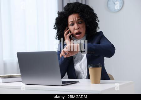 Deadline concept. Scared woman talking on smartphone and checking time in office Stock Photo