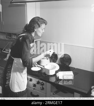 In the kitchen 1950s. Interior of a kitchen and a young woman standing at the kitchen cooker with something cooking that is is tasting. She is wearing a plastic apron.  She is Haide Göransson, 1928-2008, fashion model and actress. Sweden 1950 Kristoffersson ref AU23-1 Stock Photo