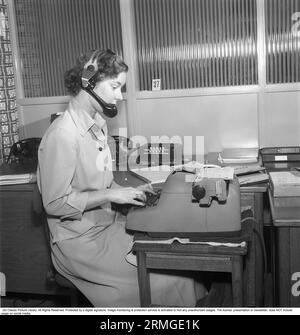 Office girl in the 1940s. A young woman sits at the typewriter and writes. She has headphones on as well as a microphone. Maybe she also manages the company's switchboard or she can also listen to a recording of someone's message and write it down. The typewriter is electric. Sweden 1946. Kristoffersson BM72-1 Stock Photo