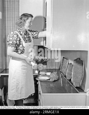 1950s home. A woman is seen preparing the table for dinner and arranges the plates and glasses she takes from a cupboard onto a tray. Sweden 1950 Stock Photo
