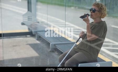 Elderly blind woman sitting at a bus stop and using a smartphone.  Stock Photo