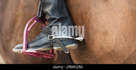 Foot of the athlete in a stirrup astride a horse, the foot of the rider, sitting on a horse, in a black boot with a spur, rests on a metal stirrup Stock Photo