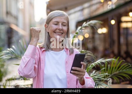 An elderly gray-haired woman walking through the evening city received an online notification of a message on her phone, a joyful pensioner holds her hand in a gesture of victory and triumph. Stock Photo