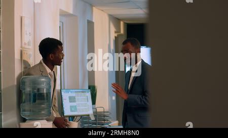 Happy coworkers discussing holiday plans during nightshift break in office. Smiling employee enjoying downtime at work, drinking cup of coffee while chatting with friendly colleague Stock Photo