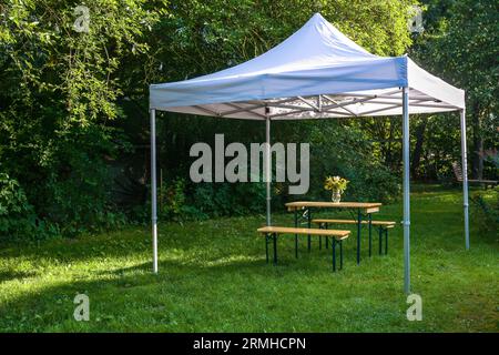 White gazebo or pavilion with a wooden table, benches and a flower bouquet on the meadow in a natural garden, idyllic summer place gives protection fr Stock Photo