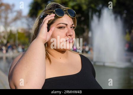 young blonde plus size latina woman standing in park at sunset upset looking at camera annoyed raising sunglasses with hands, standing with water foun Stock Photo