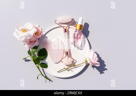 A transparent bottle with a cosmetic product with serum or rose oil lies in a plate with rose petals, a quartz massager, a gua sha scraper. Top view. Stock Photo