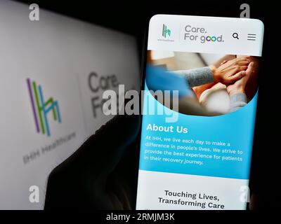 Person holding cellphone with webpage of Malaysian health company IHH Healthcare Berhad on screen with logo. Focus on center of phone display. Stock Photo