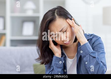 Sick woman alone at home, Asian depressed woman having severe headache, sitting on sofa in living room at home, holding hands to head from pain. Stock Photo