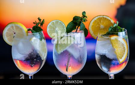 Three Colorful Gin Tonic Cocktails In Glasses At Bar Counter In Pup Or Restaurant With Out Of Focus Background Of A Sunset On A Beach Stock Photo