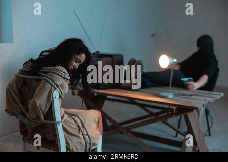 Exhausted woman sleeping and sitting on a chair tied with a rope, male kidnapper sitting opposite with a gun. Crime, kidnapping, violence concept Stock Photo