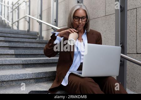 60 year old leader caucasian business lady dressed in a brown stylish suit holds a business meeting online in an urban environment Stock Photo