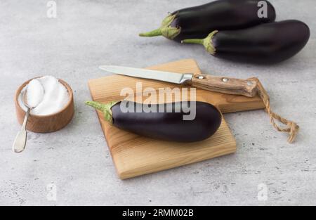 Fresh eggplant on a wooden board, a bowl of salt and a knife on a gray textured background. Cooking delicious vegan food. Stock Photo