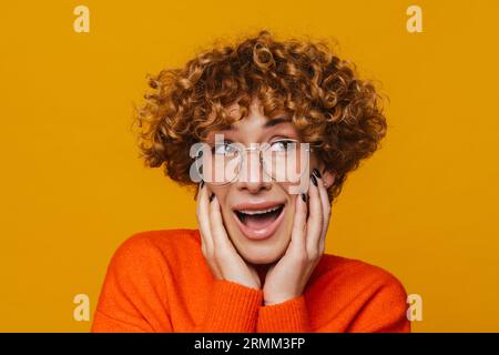 Surprised middle-aged ginger woman in glasses posing isolated over yellow studio background Stock Photo