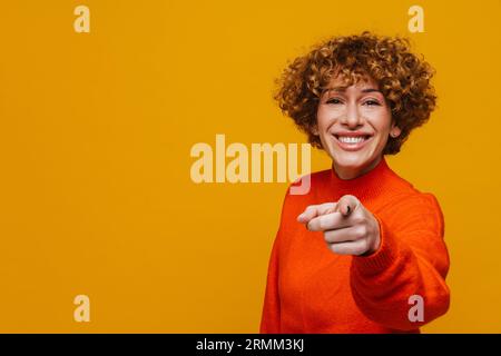 Cheerful curly middle-aged woman pointing finger at you isolated over yellow studio background Stock Photo