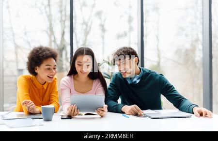 Happy diverse students using digital tablet, watching videos online sitting at table over window, copy space Stock Photo