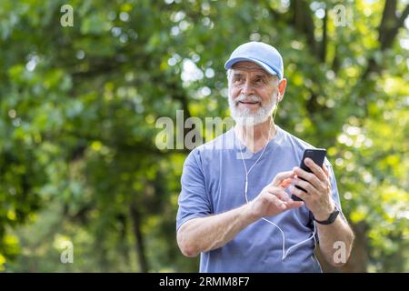 Senior gray-haired male athlete jogging in the park wearing headphones resting and listening to music online, using smartphone app, pensioner leading an active lifestyle. Stock Photo