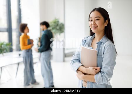 Happy asian student lady holding workbooks and smiling at camera, standing in classroom with groupmates on background Stock Photo