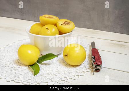 A still life with ripe yellow plums in a white porcelain bowl on a crocheted doily with one of them split by a Swiss Army knife Stock Photo