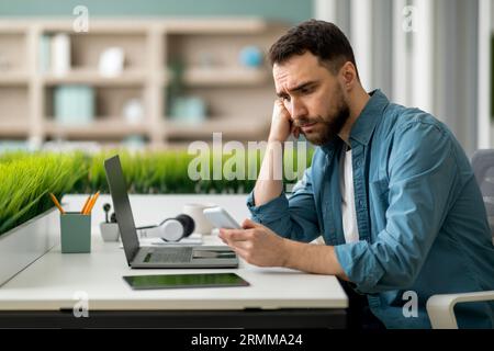 Upset Young Male Employee Using Smartphone At Workplace In Office Stock Photo