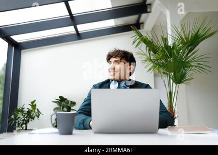 Pensive guy looking away while working on laptop, dreamy male freelancer thinking on ideas, sitting in coworking space Stock Photo