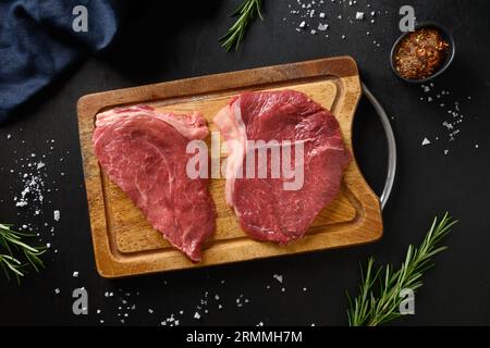 Cooking raw marbled ribeye steak with rosemary sprig on cutting board on black background. Top view. Stock Photo