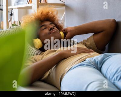 African young man lying on a sofa with eyes closed listening to music with headphones. Relaxation on the sofa Stock Photo