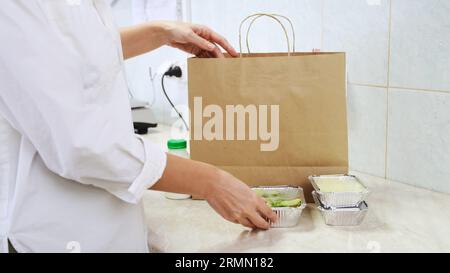 Food delivery service in restaurant. Chef packs lunch boxes to paper bag. Healthy diet daily menu, food meal dishes in a plastic takeaway containers. Woman cook on white kitchen collecting food box Stock Photo