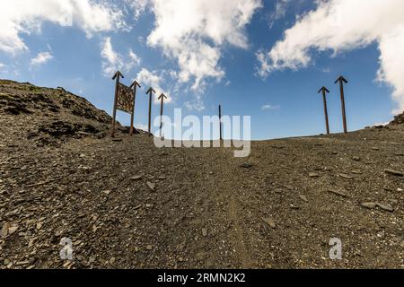 The top of the Atsunta Pass (Georgian აწუნთის უღელტეხილი) in Georgia is dotted with rusty metal posts with arrowheads. On the left, a board covered with stickers and plaques hangs between two posts. A narrow path leads up over the slate scree to the crest. White clouds drift across the blue sky above. The Atsunta Pass links the regions of Tusheti and Khevsureti. It is more than 3,400 metres high. At this altitude, plants grow for only a few weeks a year. Snow covers the ground most of the time. The semi-nomadic Tushetians live in the mountain villages only from July to September. They spend th Stock Photo