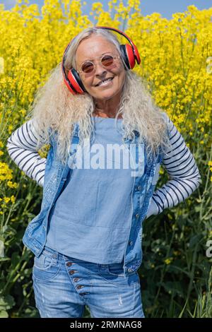Cheerful senior woman with headphones standing beneath a rapeseed field in summertime. Stock Photo