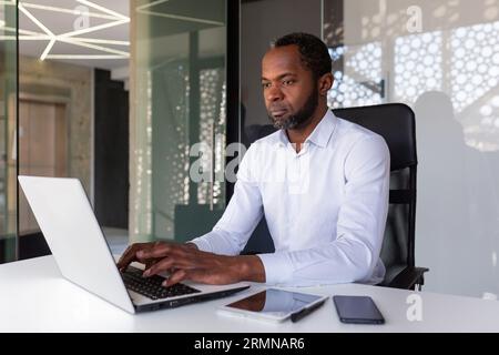 Serious focused african american programmer working inside office, man coding new software on laptop, mature experienced team leader developing technical project. Stock Photo