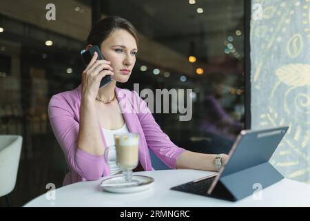 Thoughtful businesswoman talking on the phone with clients or business partners while looking at laptop monitor at cafe table Stock Photo