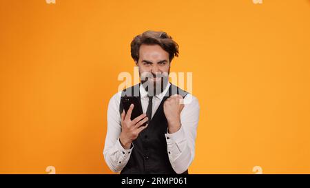 Joyful butler uses smartphone app in studio, feeling happy after reading messages on camera. Confident stylish worker with suit and tie browsing online on mobile phone, working at restaurant. Stock Photo