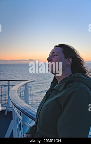 Beautiful woman relaxing against the boat balcony railing with her eyes closed and the wind in her hair. Glowing sunset on the Scandinavian sea coast Stock Photo