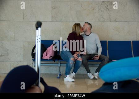 Young caucasian couple kissing while waiting, sitting on a bench at the airport Stock Photo