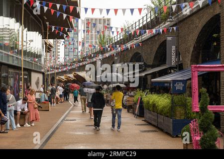 People walking in the rain and holding umbrellas on Arches Lane near Battersea Power Station next to various restaurants and bars. London, England Stock Photo