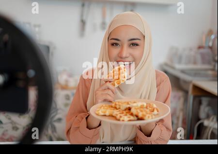 A beautiful Asian Muslim female lifestyle influencer or food blogger is eating waffles in the kitchen while recording her video or live streaming on s Stock Photo