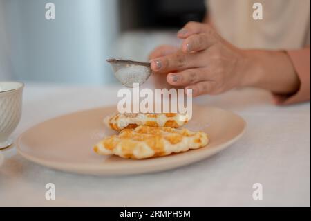 Close-up image of a female sprinkling icing sugar over waffles in a plate on a table in the kitchen. Delicious homemade pastry Stock Photo