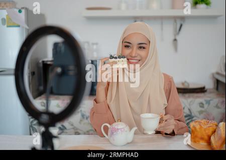 A beautiful Asian Muslim female food blogger is eating a yummy cake and hot tea at a table in the kitchen while recording her video for social media. Stock Photo
