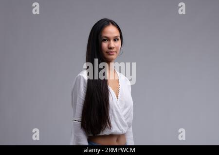 Portrait of a beautiful young woman with long black hair, wearing white blouse, blue jeans and with hoop earrings, standing against grey background an Stock Photo