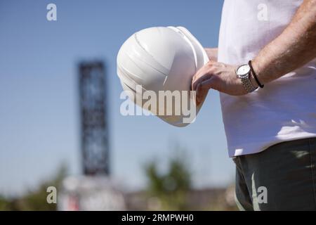 a worker in a white T-shirt holds a white construction helmet in his hands at a construction site Stock Photo
