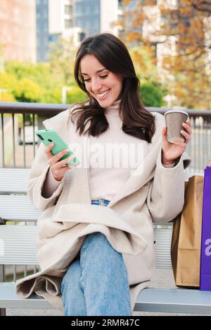 Vertical portrait of a happy young woman sitting on a bech using a smartphone to text messages after shopping on-line. Brunette female looking to the cellfone device sharing content on social media. High quality photo Stock Photo