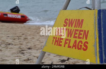 Warning sign to swim between the flags on a Queensland beach has rough sea and a surf life saving rescue inflatable boat in the background Stock Photo