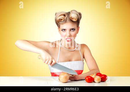 Young blonde housewife chopping onion on table in kitchen. Retro classic 50s style photoshoot. Stock Photo